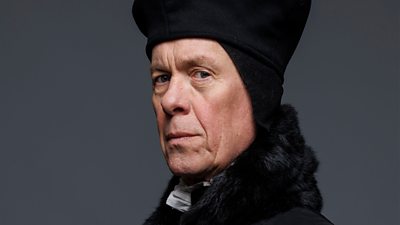 Alex Jennings in character as Stephen Gardiner. Dressed in formal clothing of the Tudor Period, he looks to the side directly to the camera. 