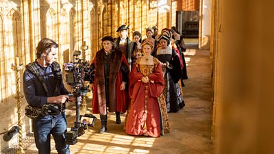 Behind-the-scenes image from Wolf Hall: The Mirror and the Light. Pairs of castmembers walk down a corridor in front of a camera operator. 