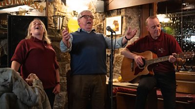 Jen (Clare Barrett); Ken Pritchard (Gregor Fisher); Ian (Robin Laing) singing in a bar with a guitar