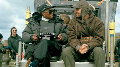 Behind the scenes shot of man and woman sitting down with filming equipment in the background