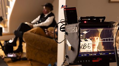 Behind the scenes shot showing Rebecca Hall in a camera screen in the foreground. in the background a blurry person sits on an armchair 