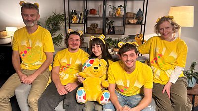 The Brockmans are sitting on a gray sofa wearing Children in Need t-shirts and Pudsey bear ear bands and holding a large Pudsey bear outfit.