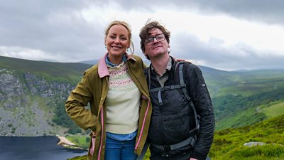 Two people with arms around each other’s shoulders smile to the camera. Dressed in hiking gear, the pair are stood on a hillside. The view behind them shows mountains, hills, and green countryside.
