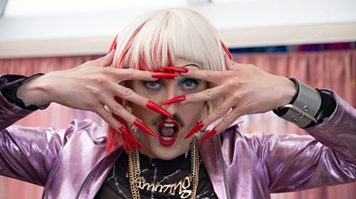 Phil Dunning as Dicke - in drag with big red nails. They've got their hands over their face and mouth open looking directly to the camera 