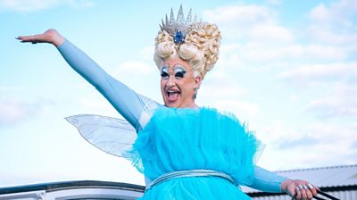 Mark Benton as Mam in full drag with a blue tulle dress, fairy wings and a crown. They're hand is up and they're smiling