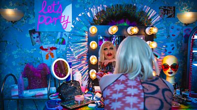 Phil DUnning as Dickie in drag looking to the camera through a lit up mirror. The background is lots of makeup and colour and peacock feathers with let's party in pink neon lights 