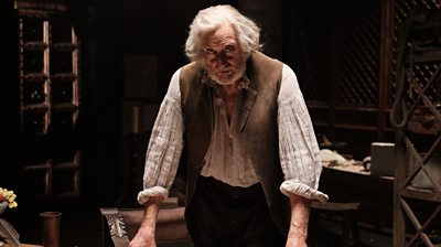 An older man stands behind a desk in an artist workshop surrounded by sculpting tools