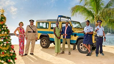 The cast of Death in Paradise stand on the beach in front of a yellow and blue striped police vehicle. A Christmas tree sits just off to the left.