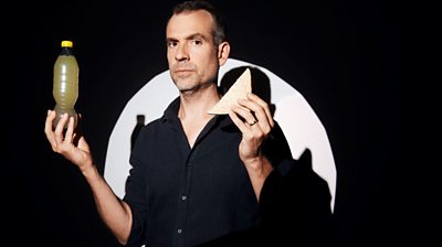 Chris van Tulleken holding up a drink of bottled juice and a sandwich and looking to the camera. Black background with a white circle behind him