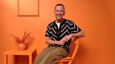 Scott Mills sits on an orange chair beside an orange table set against an orange wall