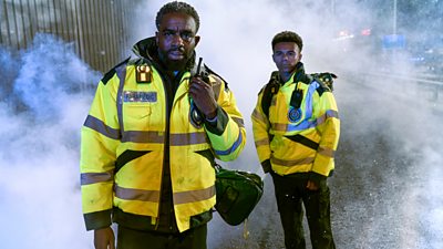 Two paramedics, played by Charles Venn and Milo Clarke, stand in high vis jackets looking concerned as cold fog swirls around them