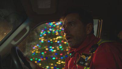 Ambulance driver Iain sits in the cab looking shocked, with a lit up Christmas tree visible through the driver's side window