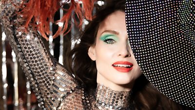 Sophie Ellis-Bextor wears a sequined outfit with dark burnt orange feathers, Only half her face is visible behind the brim of a black bejewelled hat she holds. She wears bright blue/green eyeshadow and bright red lipstick.