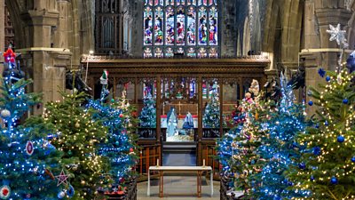 Inside of a church filled with lit up Christmas trees and a stain glass window and Nativity set in the background