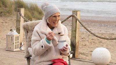 Mary Berry sits in a chair on a deck by the sand and sea, wearing warm clothes and laughing as she eats from a tin mug with a spoon