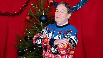 Eric Knowles wears a brightly patterned Christmas jumper and stands beside a Christmas tree