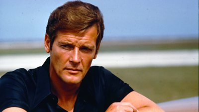 Roger Moore wears a short sleeved dark shirt and looks at something off camera