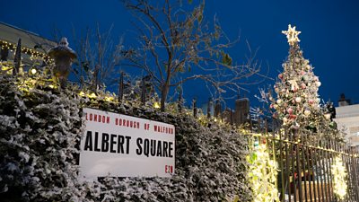 Snow covers the bushes and a Christmas tree on Albert Square, lit up by Christmas lights at night