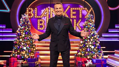 Bradley Walsh wears a dark suit and stands between two brightly decorated Christmas trees on the lit up set of Blankety Blank