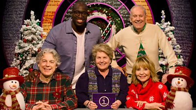 Alan Davies, Emmanuel Sonubi, Sandi Toksvig, Gyles Brandreth and Lulu smile for the camera sitting and standing behind a desk decorated with snowmen with Christmas trees in the background