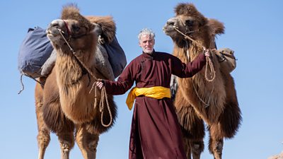 Gordon Buchanan stands in the Bumbat foothills with a camel called Chewy on the right and a camel called Squeaks on the left.