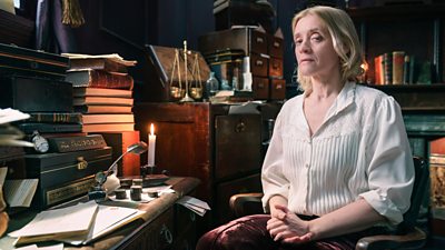 Anne Marie-Duff sits at an old writing desk, wearing a white blouse and looking solemn