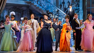 People dressed in bright gowns and suits. Hanna Glawari (Danielle De Niese), Viscount Cascada (Alex Otterburn) And Raoul De St Brioche (Innocent Masuku) with The Glyndebourne Chorus.