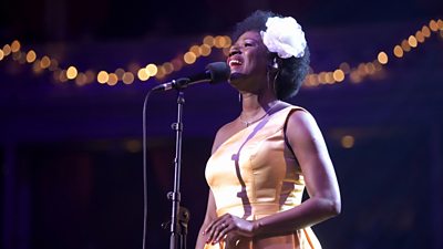 A woman sings at a microphone, wearing a gold gown with a large white flower in her hair