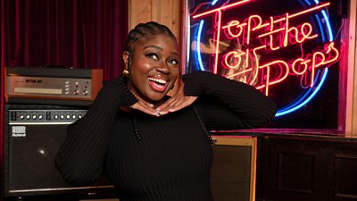 Clara Amfo smiles in front of some amps and a neon Top of The Pops sign