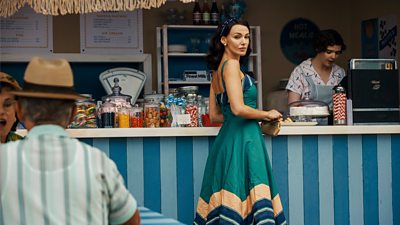 Kate Thorne (Michelle Keegan) wears a blue dress and looks over her shoulder at something off-screen. She has been standing at an outdoor food kiosk. 