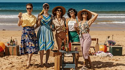 A group of women stood on the beach in 1950s fashion. All five women smile for the camera, surrounded by chairs, food and drinks for a seaside picnic in the sun.  