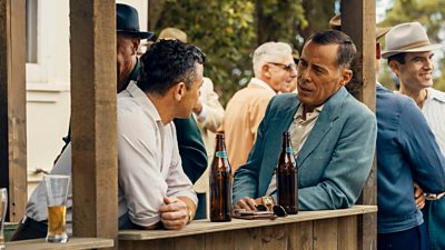 Two men lean on an outdoor wooden shelf and talk. Beer bottles, sunglasses and wallets are on the shelf. 