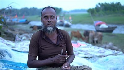 A fisherman looks at the camera as he holds a penknife and line. He is sitting among his nets with the river, some cattle and some small boats in the background.