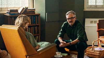 Two people are sitting opposite each other. A woman in an armchair faces away from the camera, while a man sits on a coffee table and looks worriedly at something off-screen. 