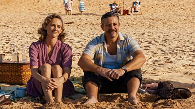 Two characters sit side by side in the sand by the beach. Their bare feet are buried in the sand and they are sitting next to a picnic blanket basket. 