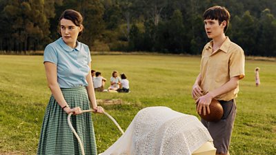 A young woman and a young man stand in a park and look at something off screen. The woman pushes a pram while the man holds a rugby ball. 