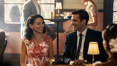 A woman and a man laugh while sitting next to each other at a table in a bar. Other patrons sit in pairs in the background. 