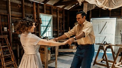 A woman and a man hold hands while dancing in a wooden warehouse. They laugh as they spin around. 