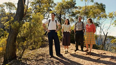 Four people stand outdoors on a hilltop. Trees and bushes surround them. A canopy of trees is visible over the edge of the hill.