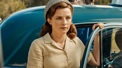 A woman holds on to the door frame of a classic car as she stares in shock at something off-screen. 