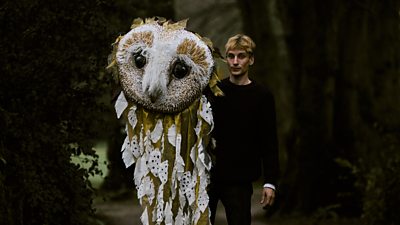 Charlie Cooper stands outdoors and holds a large, elaborately decorated craftwork with the face of an owl. The decoration trails to the floor and is as tall as Charlie. 