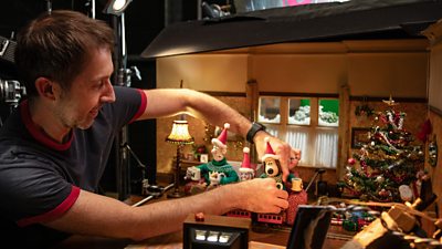 A man adjusts a figurine of Gromit the dog on a sofa in a Christmassy living room scene