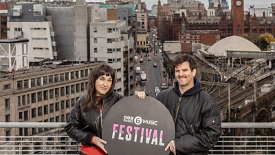 Two people hold a sign on a balcony. A city skyline is in the background. Text on the sign reads: “ý Radio 6 Music Festival.” 