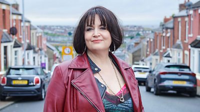 A woman smiles to the camera. She's stood on a residential street at the top of a hill. A view of the town is visible in the background.