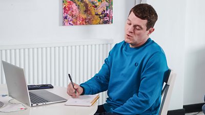 A man sits at a kitchen table and writes a list on a notebook. There’s a laptop sat on the table in front of him. 