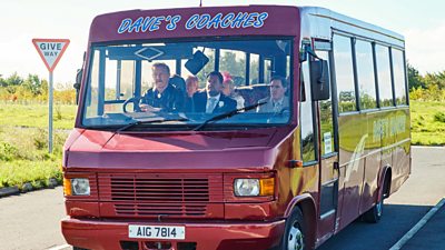 A red coach branded “Dave’s Coaches” waits on a a road near a junction. Five characters are sat on the coach, looking at something out of the windscreen. 