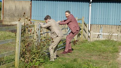 Two men wearing suits struggle to open a large, metal gate between two fields. 