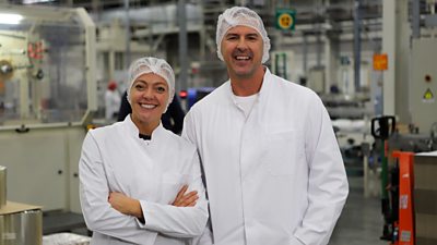 Two people stand in white overalls and hairnets on a factory floor. Large, industrial machinery is visible in the background. 