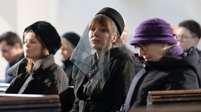 Philomena Cunk (Diane Morgan) sitting in a church dressed in black with a black veil over her face and looking at the camera