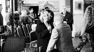 A black and white behind-the-scenes photo from filming. A woman and a man chat in between filming. Other cast and crew are visible in the background. 